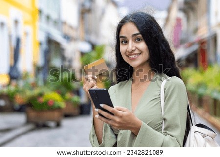 A portrait of a young beautiful Indian woman standing on the street, holding a crew and a phone in her hands, smiles at the camera. The photo is close.