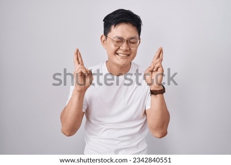 Young asian man standing over white background gesturing finger crossed smiling with hope and eyes closed. luck and superstitious concept. 