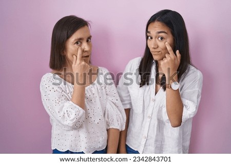 Hispanic mother and daughter together pointing to the eye watching you gesture, suspicious expression 