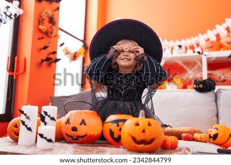 Adorable blonde girl wearing witch costume holding sweet over eyes at home