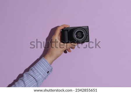 Man's hand in shirt holding modern digital camera on purple pastel background with shadow.