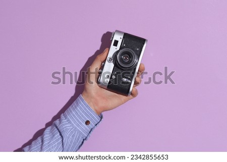 Man's hand in shirt holding retro film camera on purple pastel background with shadow.