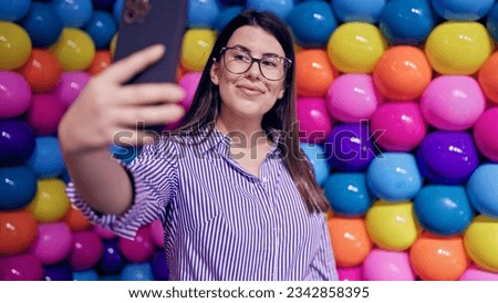 Young beautiful hispanic woman visiting colorful futuristic exhibition space taking selfie 