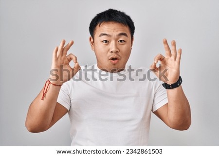 Young chinese man standing over white background looking surprised and shocked doing ok approval symbol with fingers. crazy expression 
