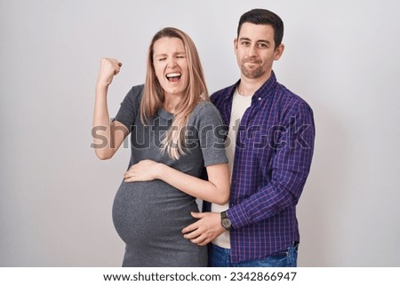 Young couple expecting a baby standing over white background very happy and excited doing winner gesture with arms raised, smiling and screaming for success. celebration concept. 