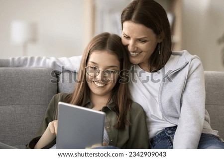 Cheerful pretty young mom and schoolgirl daughter in glasses using online service, educational application on tablet computer gadget, resting on home couch, shopping on Internet Royalty-Free Stock Photo #2342909043