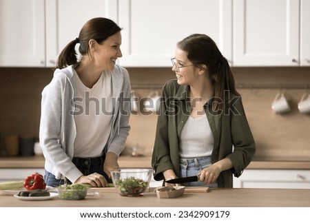 Cheerful mom and positive teenage daughter cooking dinner in home kitchen, preparing salad, slicing from fresh vegetables, talking, laughing, enjoying culinary hobby, family domestic activities Royalty-Free Stock Photo #2342909179