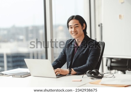 Portrait of handsome asian businessman, successful trader, using laptop, working in modern office, looking at camera. Freelancer copywriter writing on keyboard, planning project sitting at workplace