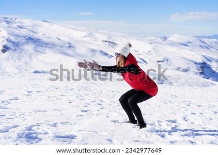 Side view of young female in warm clothes cap sunglasses looking away while squatting, with stretched hands on snow against blue sky and snowy mountains in day light
