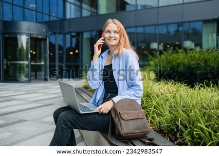 Young Caucasian student freelancer 30s woman rest use laptop pc computer talk by mobile cell phone look, outdoors sits against blurred city building. She is keyboarding on laptop. Freelance work