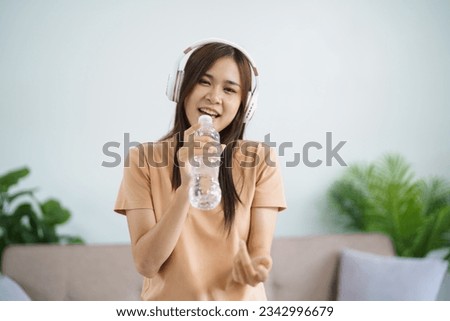 Happy Young Asian woman singing alone with bottle like microphone and wearing headphone love music happy relax in living room.