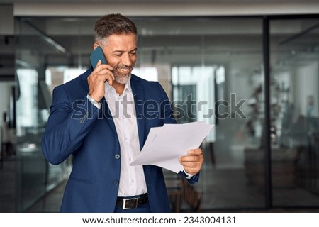 Middle aged Latin or Indian businessman walking, having call on smartphone with business partner and looking at documents. Mature Hispanic man talking by mobile cellphone at work in office, copy space
