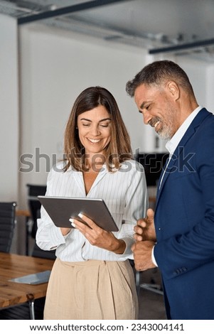 Vertical portrait of mature Latin businessman and European businesswoman discussing project on tablet in office. Two diverse partners colleagues of confident professional businesspeople work together. Royalty-Free Stock Photo #2343004141