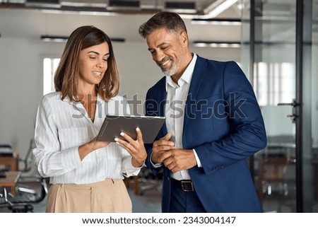 Two diverse partners walking mature Latin business man and European business woman discussing project on tablet in office. Two colleagues of professional business people working together at workspace. Royalty-Free Stock Photo #2343004147