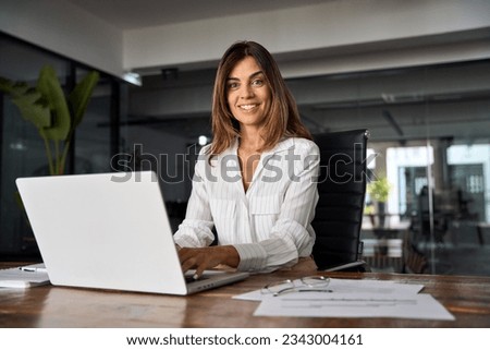 40s mid age European business woman CEO using laptop for work sitting at table in office and looking at camera. Smiling Latin Hispanic mature adult professional businesswoman using pc digital computer