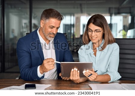Mature 50s age Latin business man mentoring mid age European business woman discussing project on tablet in office. Two colleagues, group of partners, professional business people working together. Royalty-Free Stock Photo #2343004191