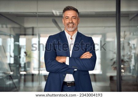 Handsome hispanic senior business man with crossed arms smiling at camera. Indian or latin confident mature good looking middle age leader male businessman on blur office background with copy space.