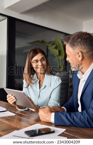 Vertical shot of team of diverse partners mature Latin business man and European business woman discussing project at table in office. Two colleagues of professional business people working on tablet.