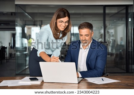 Team of diverse partners mature Latin business man and European business woman discussing project on laptop sitting at table in office. Two colleagues of professional business people working together. Royalty-Free Stock Photo #2343004223