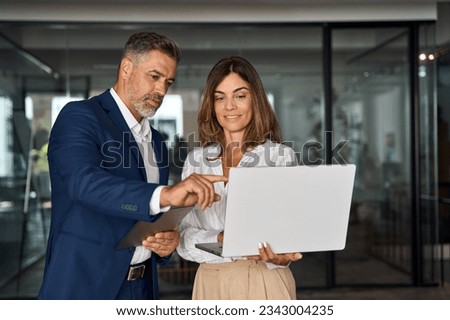 Mature Latin business man and European business woman discussing project on laptop gadget in office. Two diverse partners, colleagues, team of confident professional business people work together. Royalty-Free Stock Photo #2343004235