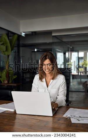 Vertical shot of European business woman CEO using laptop application sitting at workplace at table in office. Smiling Latin Hispanic mature adult professional businesswoman using pc digital computer. Royalty-Free Stock Photo #2343004241