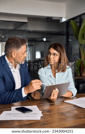 Mature 50s age Latin business man mentoring mid age European business woman discussing project on tablet in office. Two colleagues, group of professional business people working together, vertical. Royalty-Free Stock Photo #2343004251