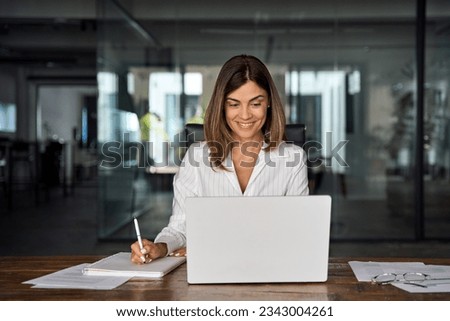 40s mid age European business woman CEO working on laptop sitting at table workspace, making notes in office. Smiling Latin Hispanic mature adult professional businesswoman using pc digital computer Royalty-Free Stock Photo #2343004261