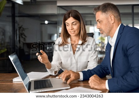 Mature 50s age Latin business man mentoring mid age European business woman discussing project on laptop in office. Two colleagues, group of partners, professional business people working together. Royalty-Free Stock Photo #2343004263