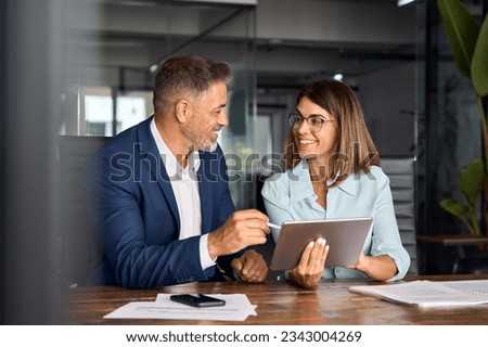 Team of diverse partners mature Latin business man and European business woman discussing project on tablet sitting at table in office. Two colleagues of professional business people working together. Royalty-Free Stock Photo #2343004269