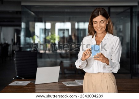 Smiling Latin Hispanic mature adult professional business woman using mobile phone cellphone. European businesswoman CEO holding smartphone using fintech application standing at workplace in office. Royalty-Free Stock Photo #2343004289