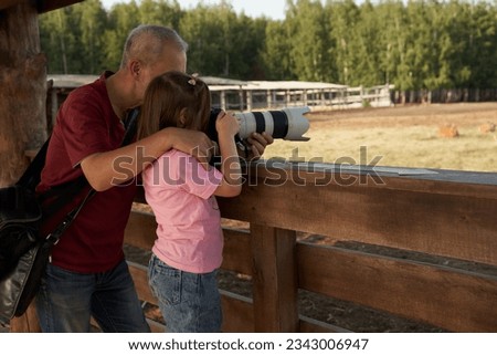 A grown man is teaching his granddaughter how to take pictures with a long lens. They are at a deer farm near a fence. The man is supporting the lens and giving explanations to the girl.  