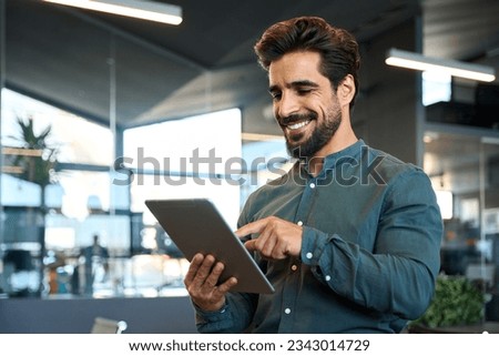 Happy young Latin business man executive holding pad computer at work. Male professional employee using digital tablet fintech device standing in office checking financial online market data. Royalty-Free Stock Photo #2343014729