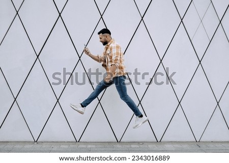A young Latino man leaps with enthusiasm, mobile phone in hand, next to a wall adorned with futuristic lines. 