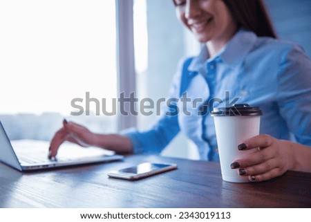 Cropped image of beautiful young business woman using a laptop, holding a cup of coffee and smiling while working in office