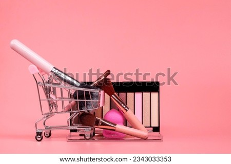 Creative concept with shopping trolley with makeup on a pink background. Perfume, sponge, brush, mascara, eye shadow, lip gloss in the basket.