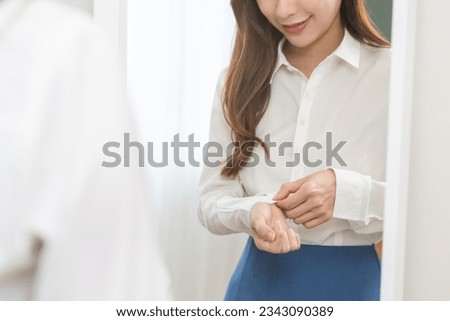 Get dress, asian young woman, businesswoman standing, hand buttoning up shirt formal, female getting dressed, ready before go to work looking reflection the mirror in bedroom in the morning at home. Royalty-Free Stock Photo #2343090389