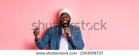 Handsome african-american man singing karaoke, reading lyrics on smartphone app and holding microphone, standing over pink background. Royalty-Free Stock Photo #2343098737