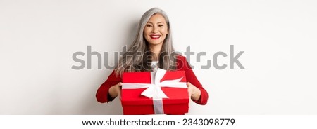 Elegant senior woman giving you present. Asian lady holding red gift box and smiling, standing over white background. Royalty-Free Stock Photo #2343098779