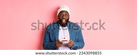 Excited Black man celebrating birthday, staring at camera amazed while making wish on bday cake with candle, standing over pink background.