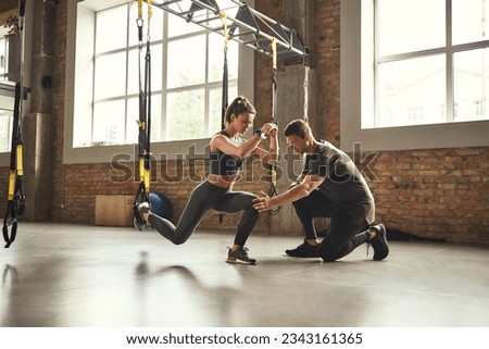 Doing squat exercise. Confident young personal trainer is showing slim athletic woman how to do squats with Trx fitness straps while training at gym. Royalty-Free Stock Photo #2343161365