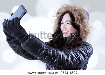 Cheerful girl wearing winter jacket with fur hood, taking self portrait using a smartphone against light glitter background