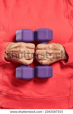 Wrinkled hands of old person holding violet small dumbbells. Healthy and fitness senior lifestyle. Senior lady exercising with weights. Shallow depth of field