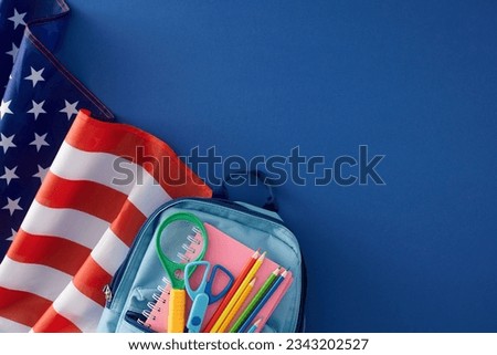 Schooltime essentials in USA concept. Top view shot of american flag, blue backpack, copybook, magnifier, scissors, colorful pencils on blue background with empty space for advert or text