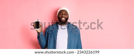Handsome hipster guy in beanie and denim shirt smiling, showing mobile phone screen with happy face, introduce application, standing over pink background.