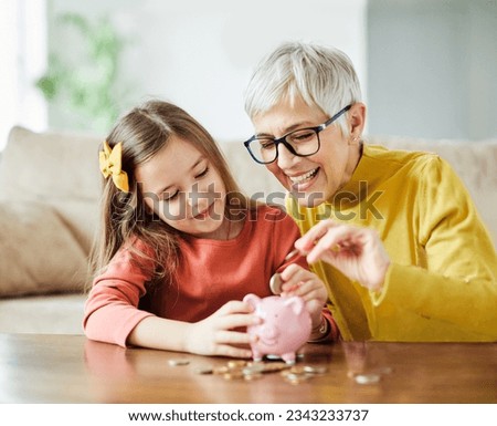 Grandmother and her granddaughter Putting Coin Money In Piggybank  At Home. Personal Savings, Bank Safety And Financial Investments Concept Royalty-Free Stock Photo #2343233737