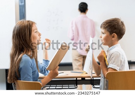 Portrait of schoolchildren sitting in a classroom and talking to each other. The schoolgirl is holding a book and dictating, the boy is holding a paper, a pencil and is ready to write. Royalty-Free Stock Photo #2343275147