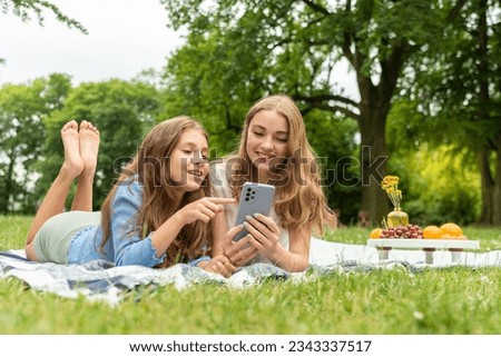 Two cute blonde girls lie in the park on a blanket on the grass, holding the phone in hands, browsing the news feed. Girls on a picnic chatting, touching the phone screen, scrolling and swiping