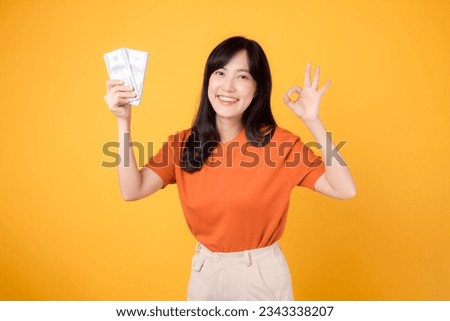 Lively young Asian woman 30s, pointing finger to cash money dollars, okay hand sign. standing on vibrant yellow backdrop.