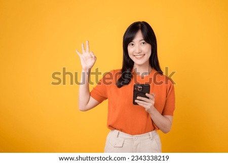 Joyful young Asian woman 30s, wearing orange shirt, using smartphone with okay hand sign on vibrant yellow background. New mobile app concept