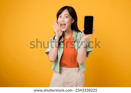 Happy young 30s Asian woman, wearing orange shirt and green jumper, presents smartphone screen with shout mouth on yellow background. New mobile application, new mobile device concept.